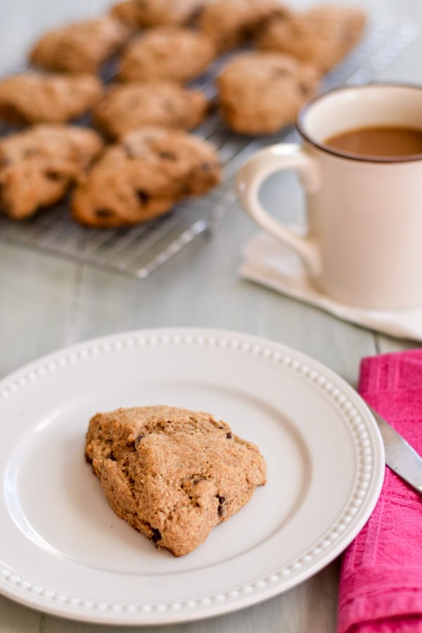 A white plate with a scone on it. There are more scones in the background with a mug of coffee to the side