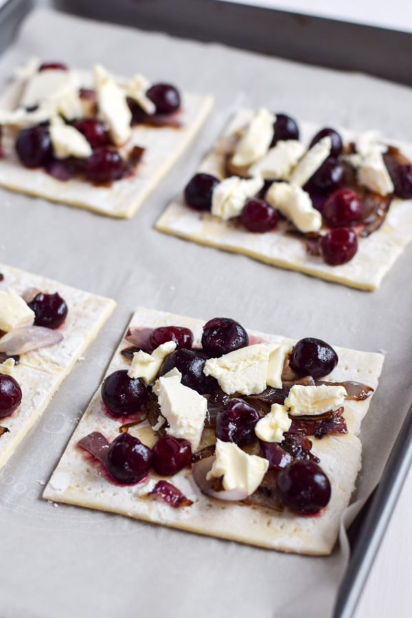 A pan with the puff pastry squares with onion, cherries and chunks of cheese on them