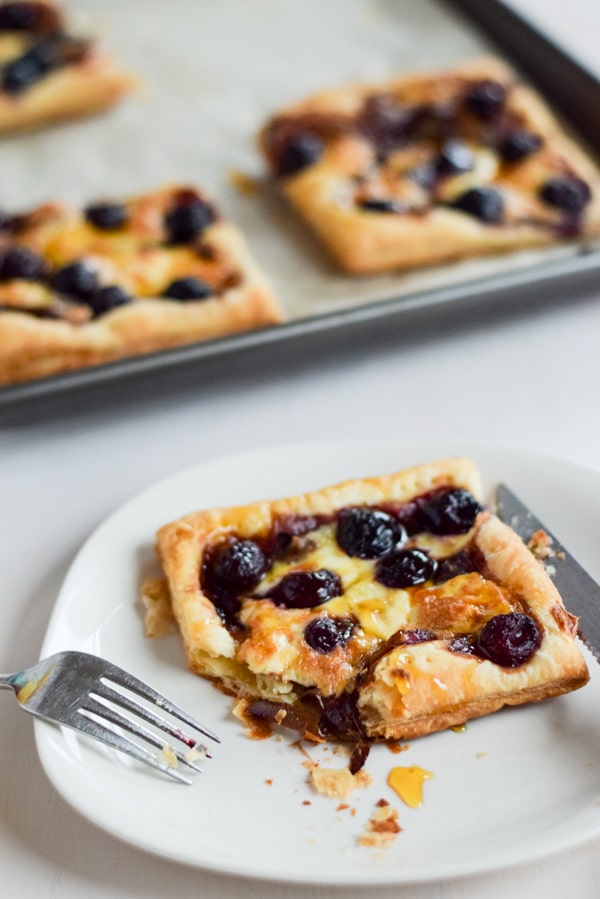 A white plate with flakey puff pastry covered with cherries, onion and cheese in front of the pan of tarts
