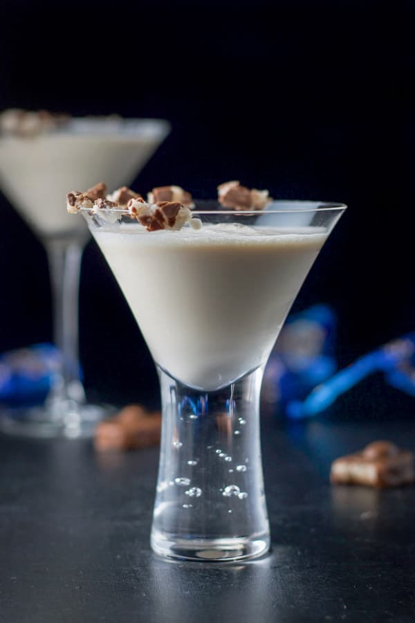 Vertical shot of the bubble glass with the milky cocktail with the candy bar on the side of the glass. There are more candy bars on the table with wrappers