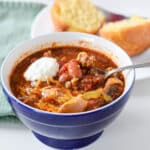 A blue bowl with the chili in it with a dollop of sour cream. There is a corn muffin on a plate - square