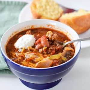 A blue bowl with the chili in it with a dollop of sour cream. There is a corn muffin on a plate - square