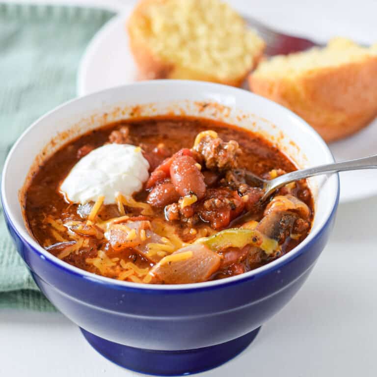 A blue bowl with the chili in it with a dollop of sour cream. There is a corn muffin on a plate - square
