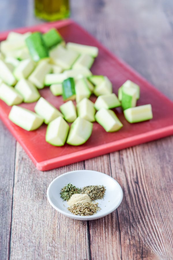 Herbs and spices in a white plate and cut up zucchini on a red board