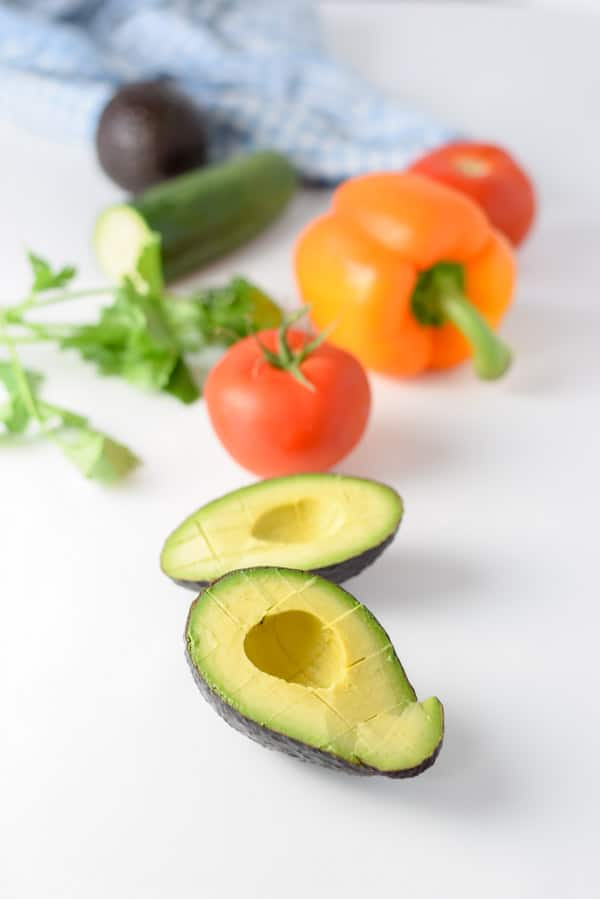 Avocado cut in half and then cubed in the skin, tomatoes, bell pepper, cilantro and cucumber on a table