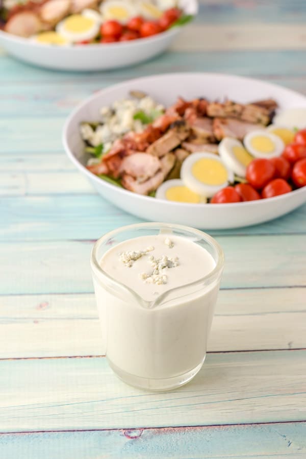 Dressing with blue cheese crumbled on top in a clear glass pitcher with two salads in the background