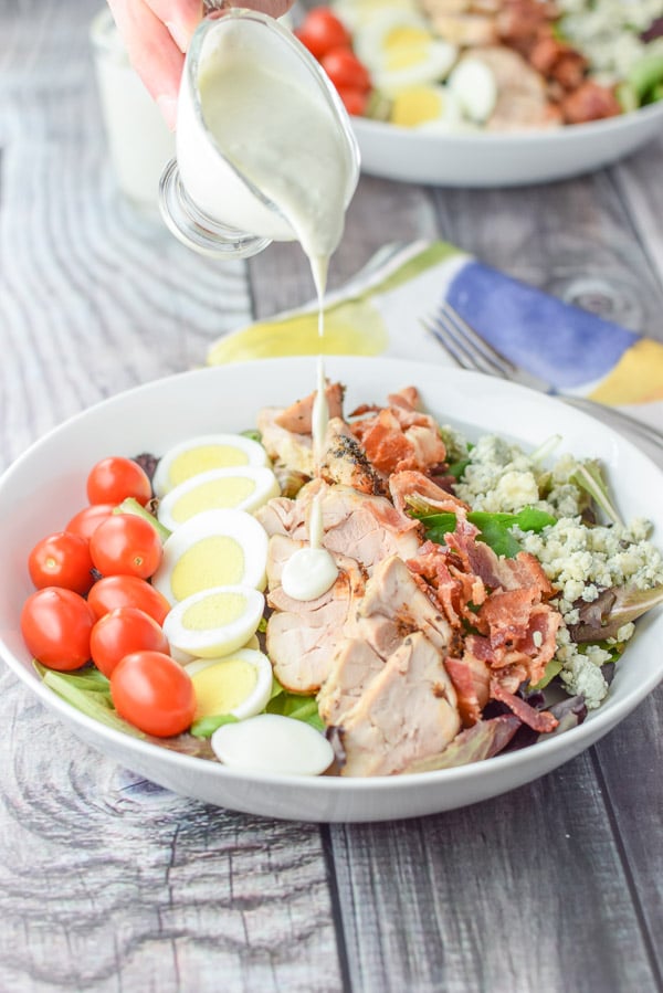 A male hand holding a pitcher of dressing pouring it on this salad with chicken, bacon egg, tomato and cheese