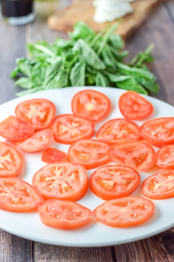 A white plate with tomato slices with basil, sliced cheese and vinegar in the background