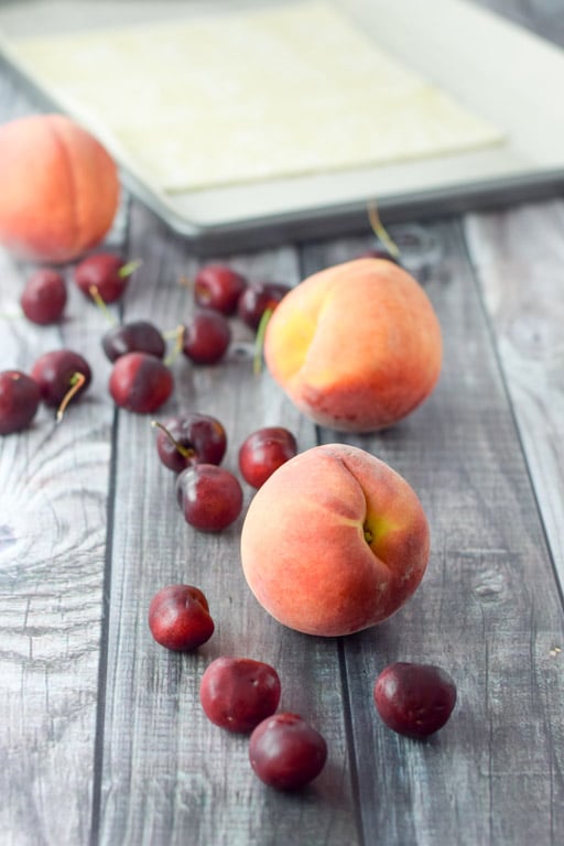 cherries and peaches in front of a pan lined with parchment paper and puff pastry on it