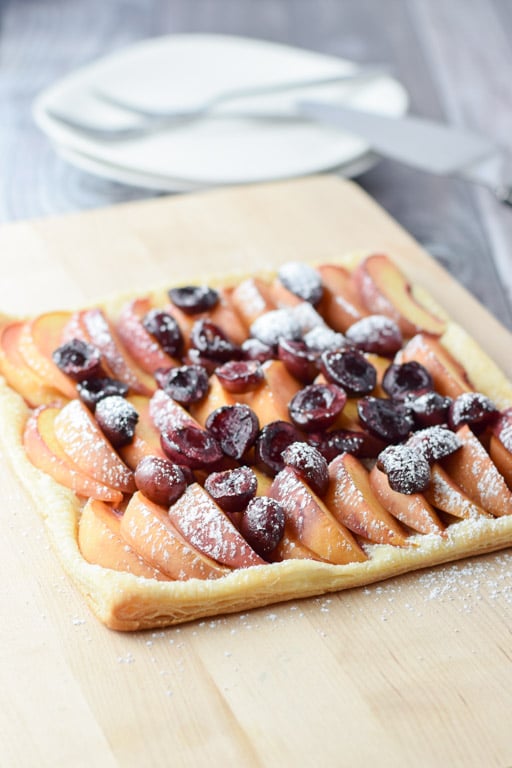 A wooden board with a fruit tart with two plates in the background