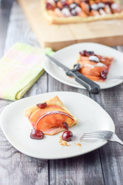 A bite taken out of a tart with cherry and peaches on it. With another plate and the tart in the background