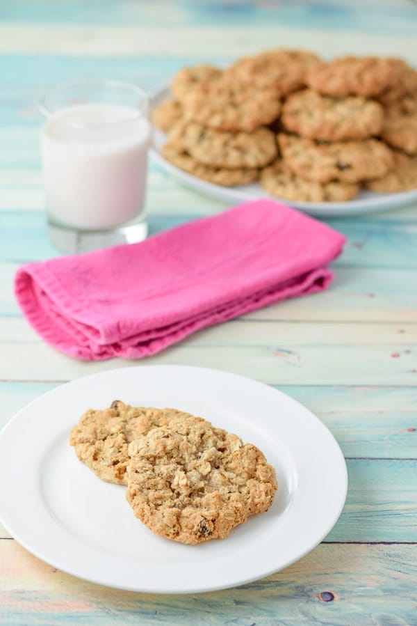 Two of the cookies on a white plate, with a glass of milk, napkin and big plate of cookies in the background