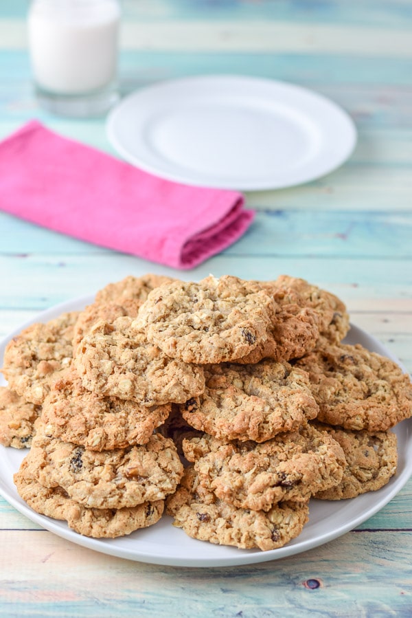 A plate piled with oatmeal cookies with a napkin, plate and milk in the background