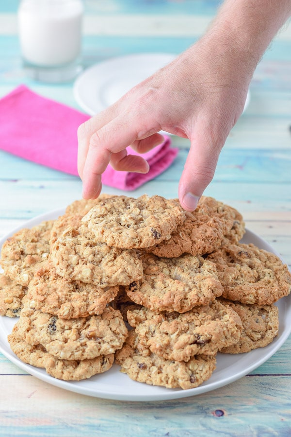 A male hand stealing a cookie off a big stack of them. There is a glass of milk, plate and napkin in the background