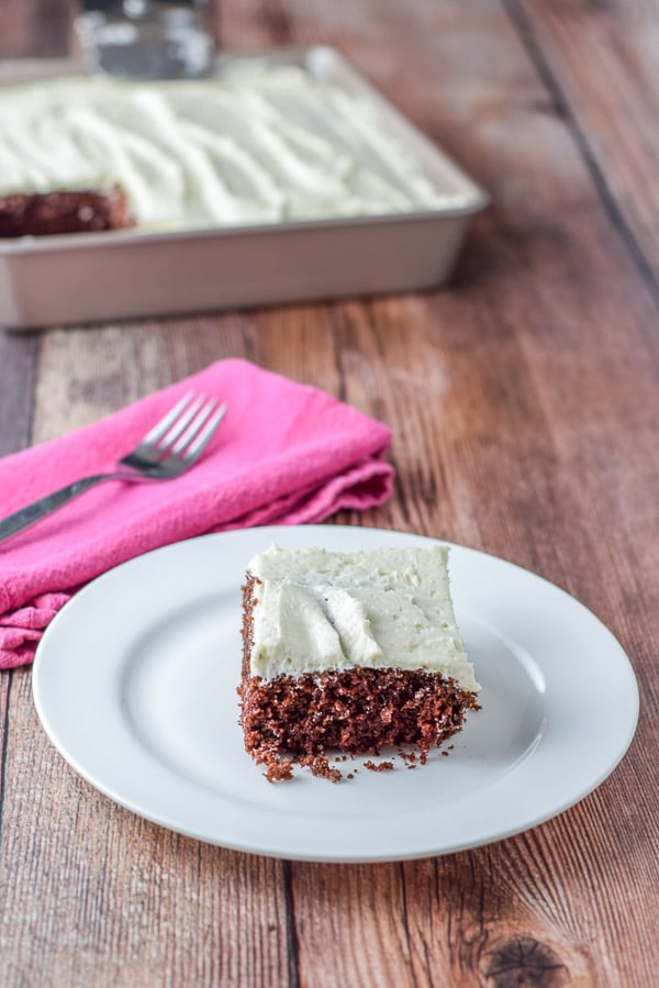 A piece of chocolate cake on a plate with a pink napkin, fork and pan of the cake in the background