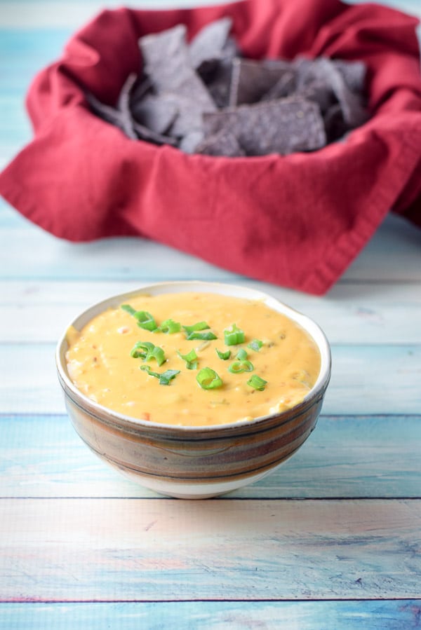 A brown bowl of cheese dip with chives and a napkin covered bowl with chips in it in the background