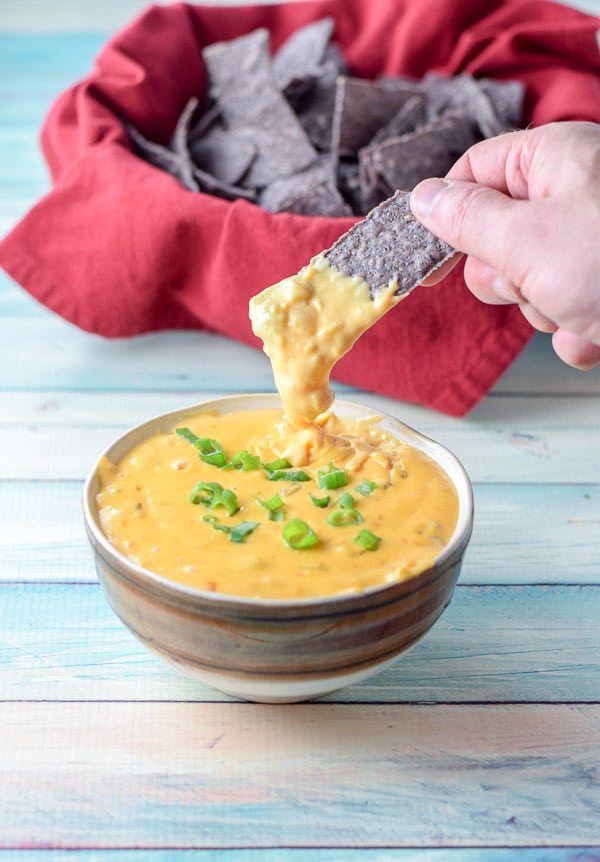 A chip dripping with cheesy queso dip and the bowl of chips in the background