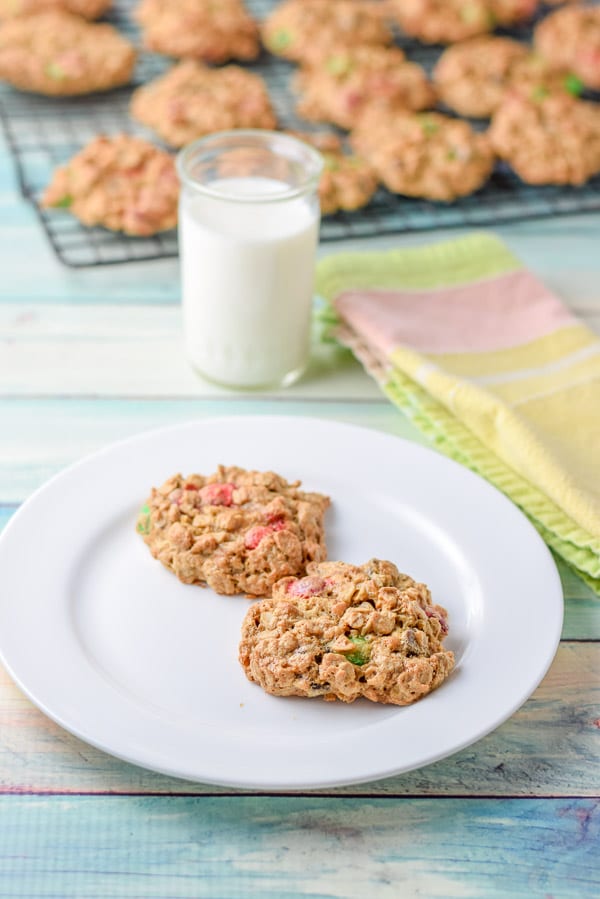 A white plate with two cookies. There is a glass of milk and a wire rack with more cookies in the background