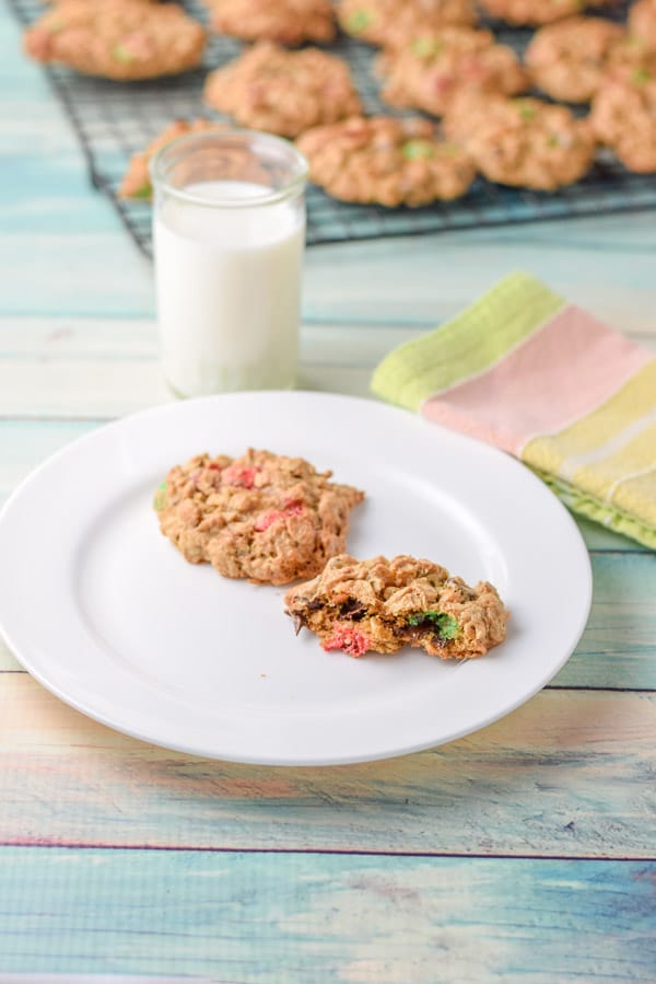 Bites taken out of the cookie with M & M's in it on a plate with another cookie, with a glass of milk and rack of cookies in the background