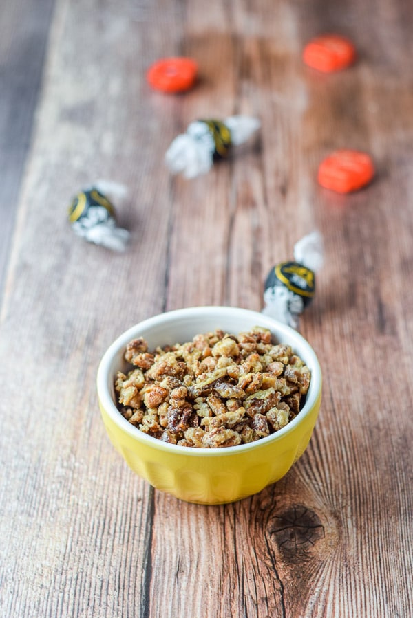 A yellow bowl filled with the candied pecans with some chocolate strewn on the wooden table