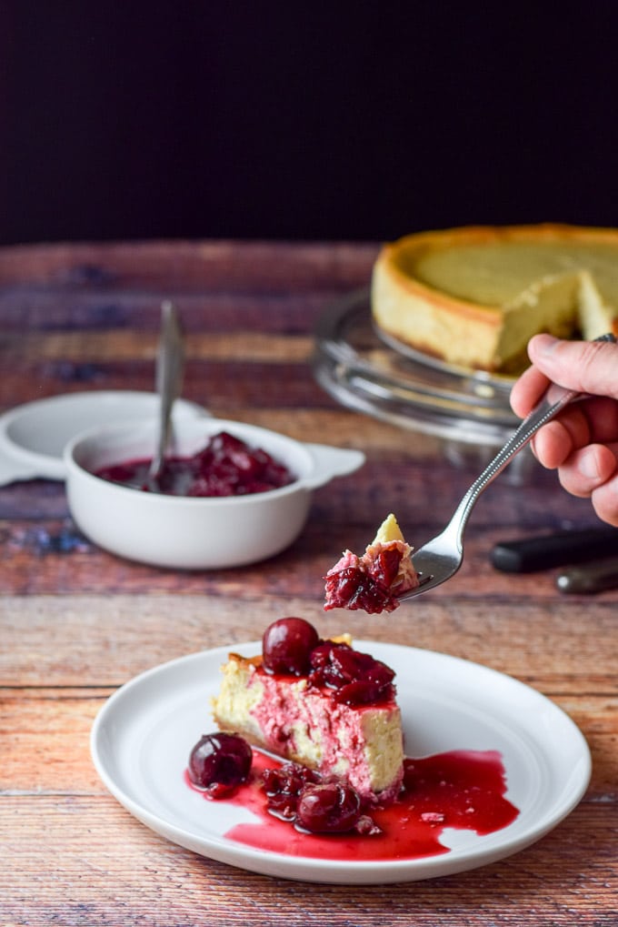 A hand holding a fork with a bite of cheesecake on it. It is held over the plate