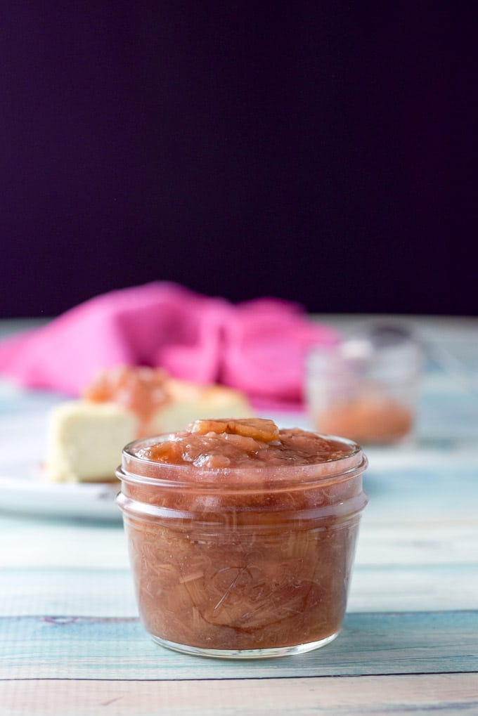 A vertical view of a one cup Ball jar full of rhubarb spread in front of cheesecake