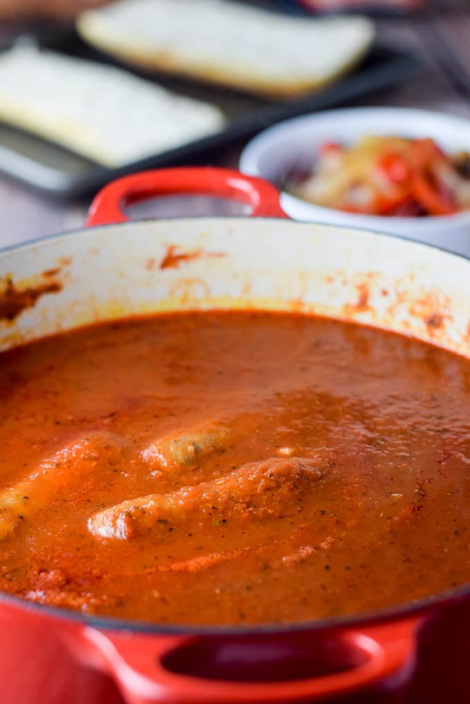 Marinara cooked with sausages in it with a bowl of cooked onions and peppers and bread on a pan in the background