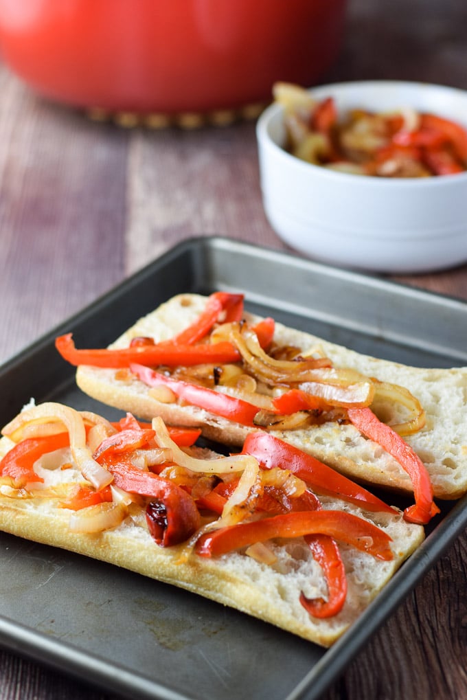 Onion and pepper on a sub roll on the pan with the veggies in a bowl behind it