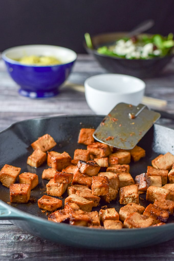 A blue cast iron pan with tofu that has been sautéed. There are some noodles and a salad in the background