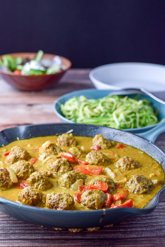 A sauté pan filled with curry sauce and meatballs. There is a bowl of zoodles and a salad in the background