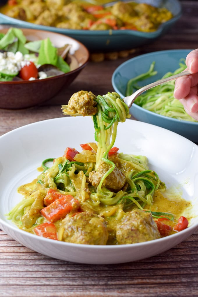 Forkful of twirled zoodles and a piece of meatball. There is a salad and the pan of meatballs in the background