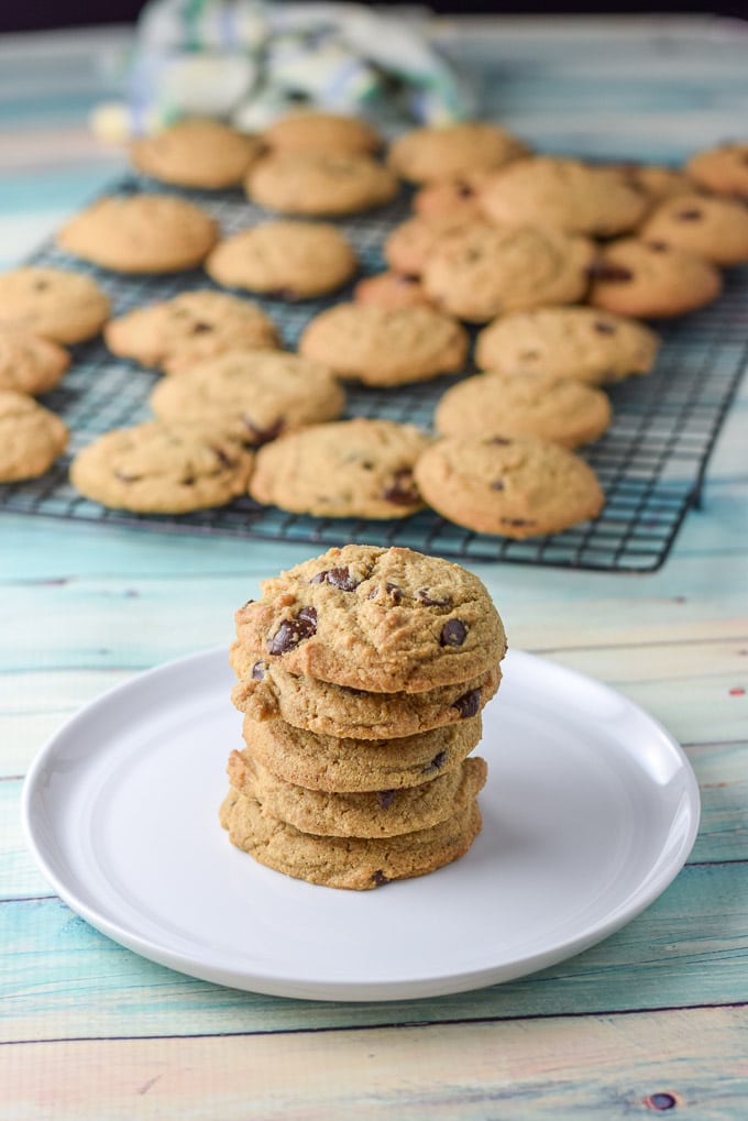 Five cookies on a small white plate piled up. There is a wire rack full of the cookies in the background