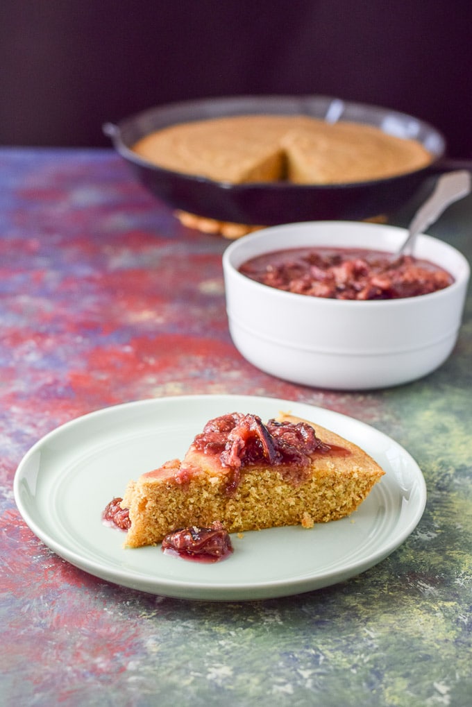 A red sauce draped over a slice of cornbread. There is a bowl of the sauce and a skillet of bread in the background