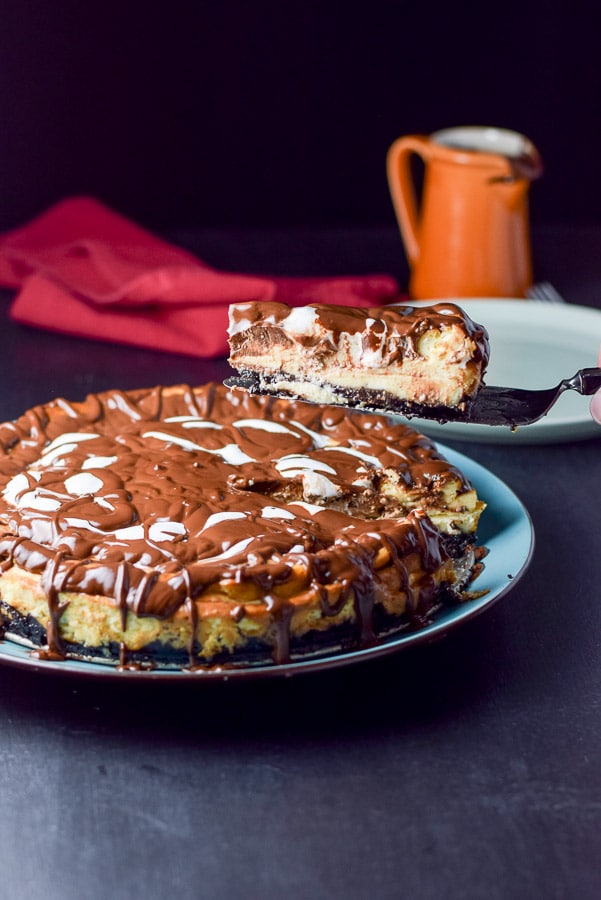 Piece of cheesecake being lifted over the cheese cake. there is a plate, fork and little pitcher of ganache in the background