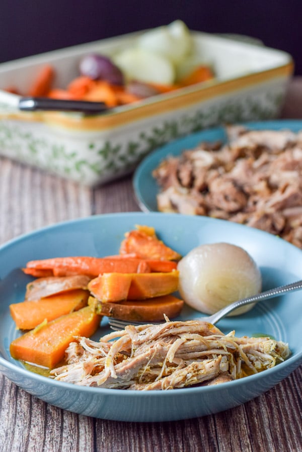 The shredded pork plated with baked vegetables. there is also a platter of pork in the background along with a baking dish of veggies