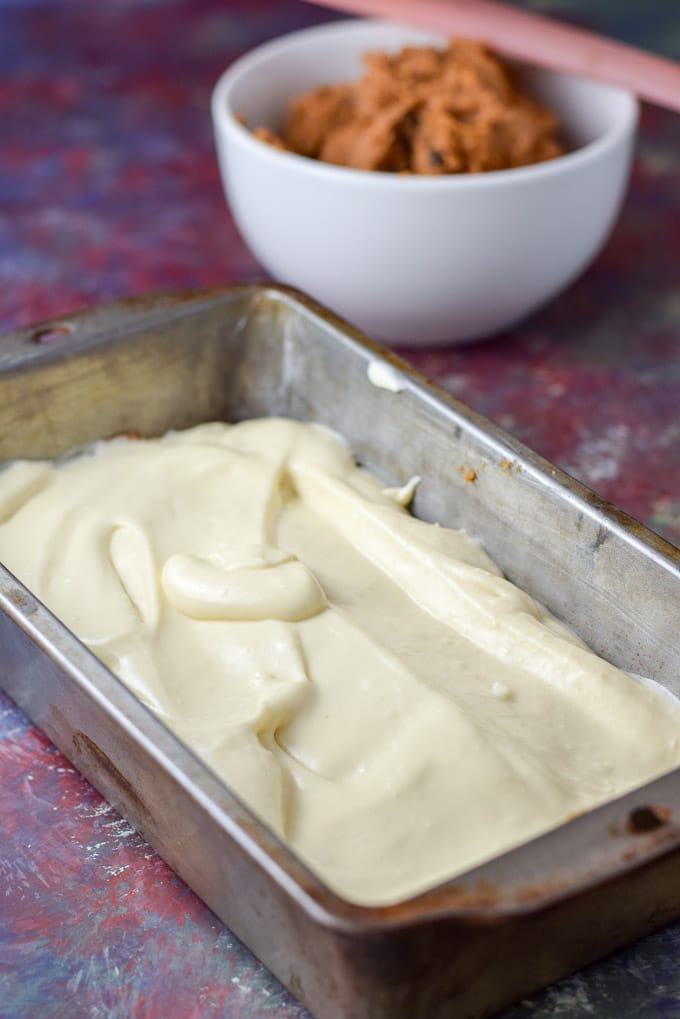 Cheesecake mixture spread on the pumpkin layer in the pan. There is a bowl of butter in the background