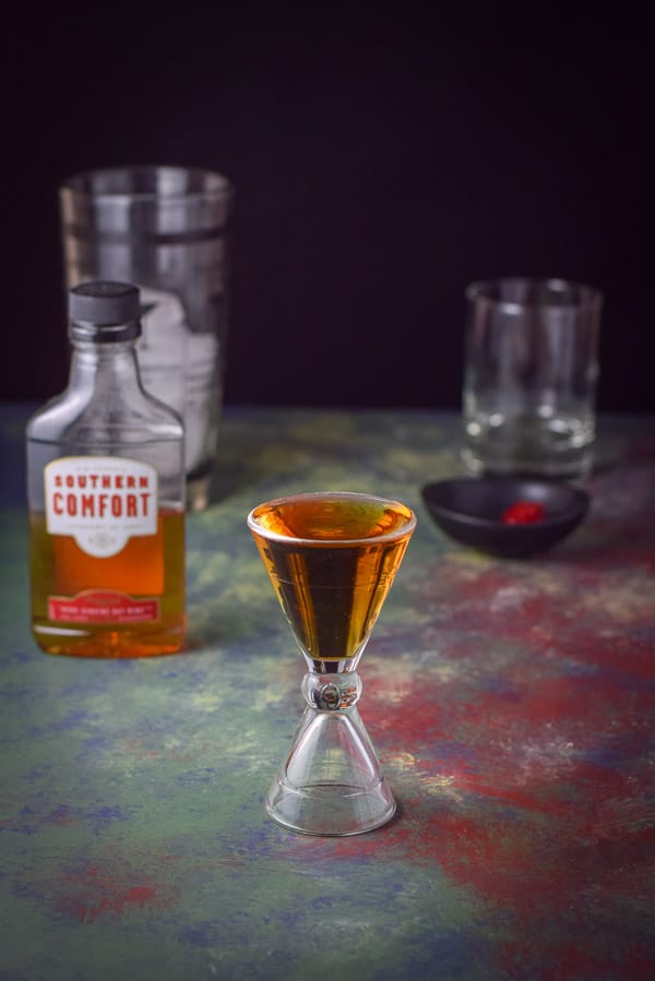 Southern Comfort poured out in a hour glass jigger with the bottle, shaker and glass in the background