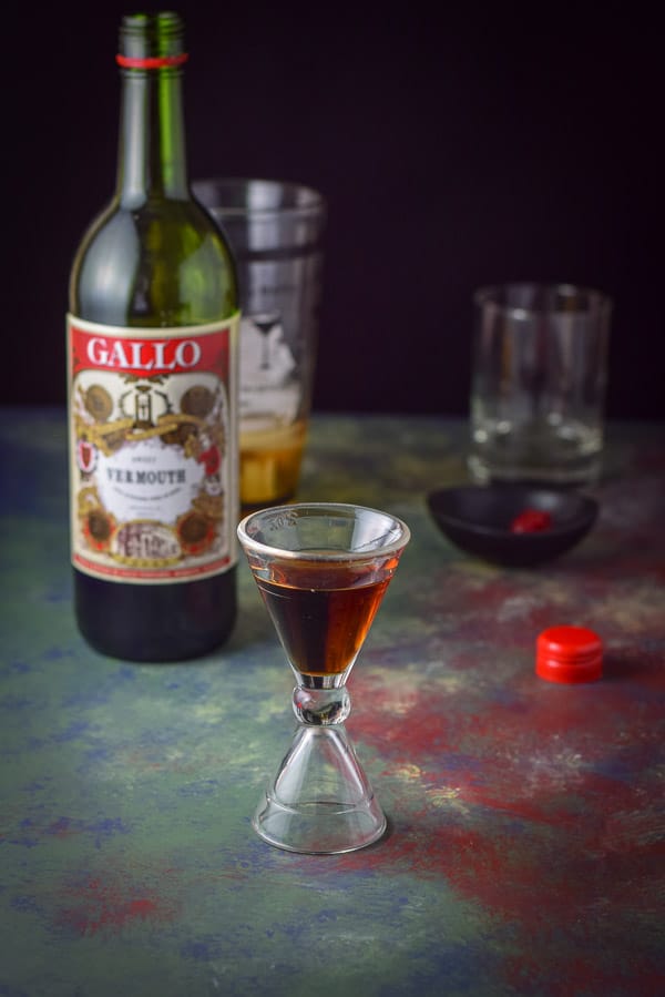 Sweet vermouth poured out in the jigger with the bottle, shaker, garnish and glass in the background