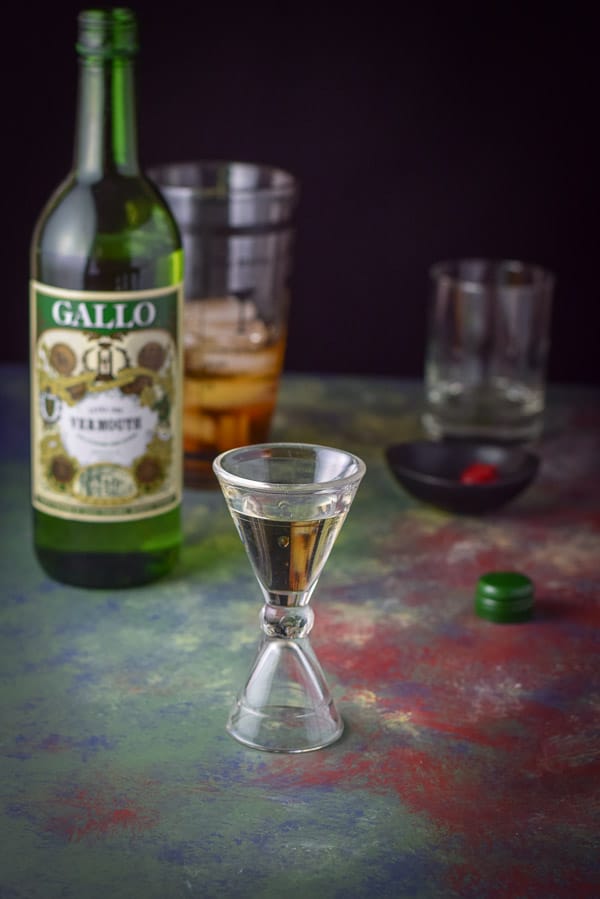 Dry vermouth measured in a glass jigger with the bottle, garnish, shaker and glass in the background