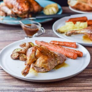 Plated leg quarter of the duck with roasted carrots and potatoes on a plate with the duck in the background - square