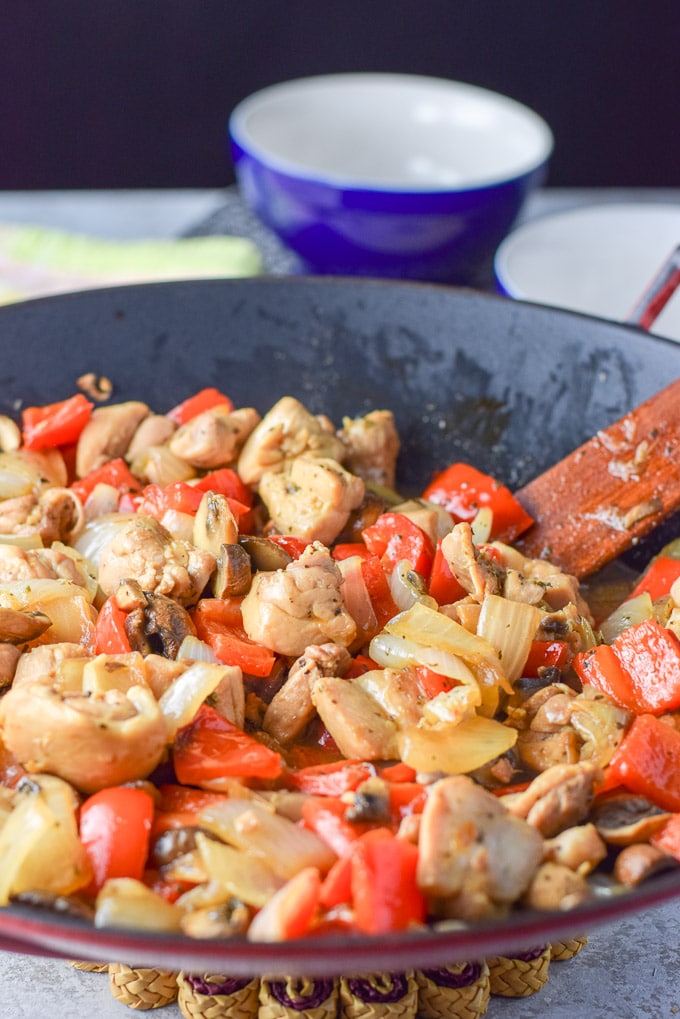 The chicken mixed with the veggies in the wok with some bowls in the background