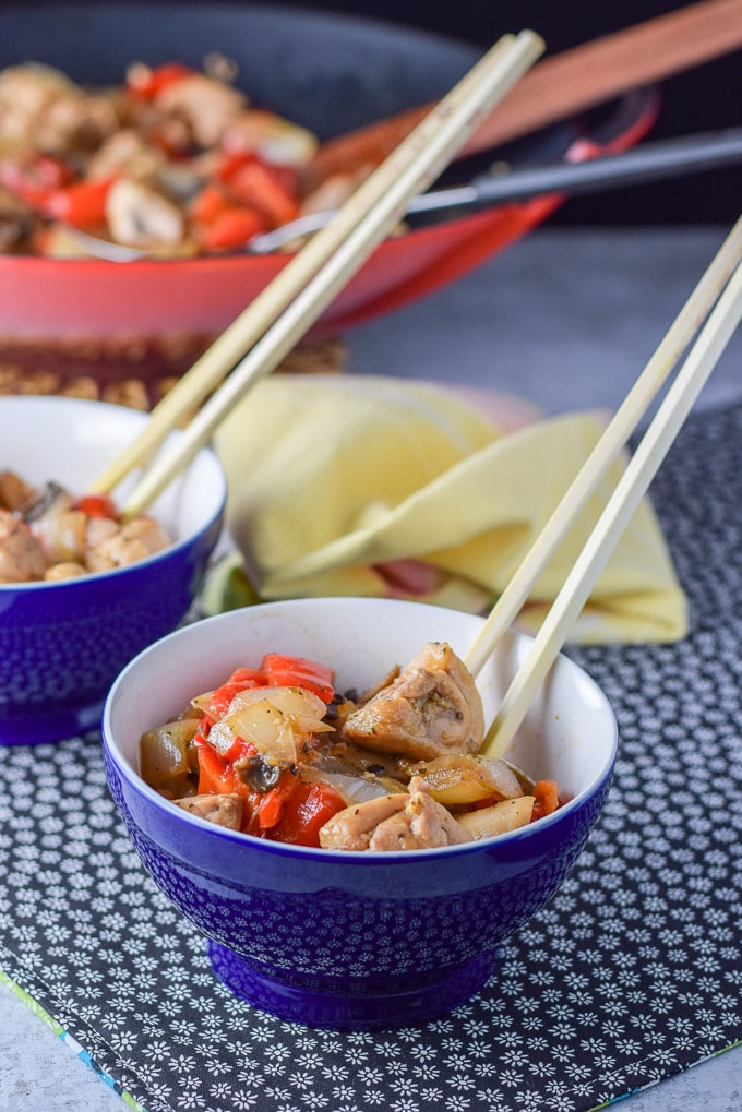 Bowl of stir fry in a bowl with chop sticks in the bowl. There is a wok filled with the dish in the background