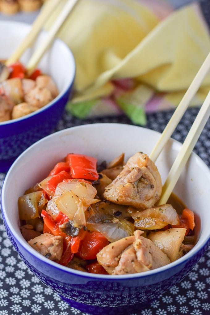 Close up of chop sticks holding a piece of chicken. There are veggies and sauce in the two blue bowls