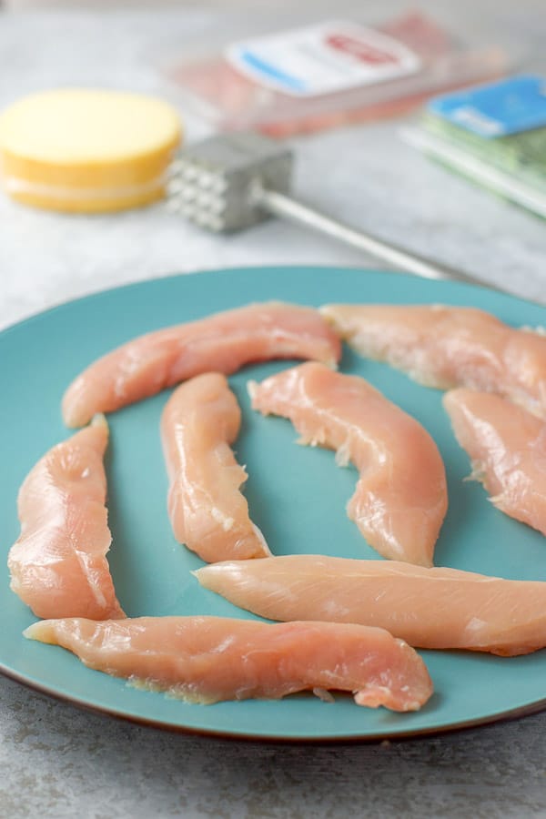 Raw chicken tenders on a blue plate, with gouda cheese, tarragon and meat in the background