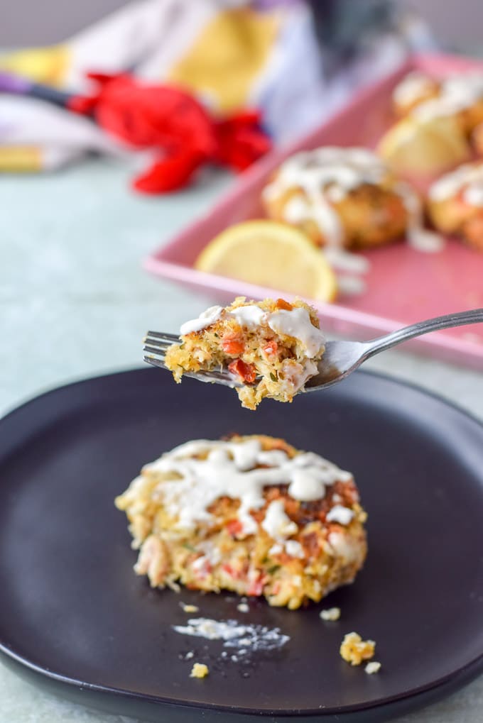 A crab cake, drizzled with sauce, on a plate with a forkful of the cake held over the plate and a platter of crab cakes in the background.