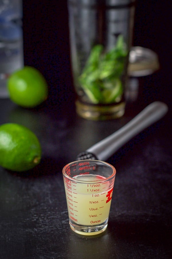 Lime juice poured out with limes in the background and mint in a cocktail shaker