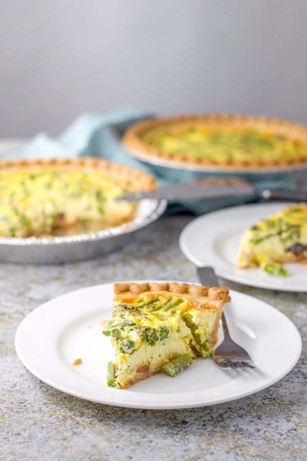 Two white plates with a piece of quiche on it with the two pies in the background