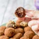A hand holding a truffle with a bite taken out of it. There is a dish of truffles underneath it