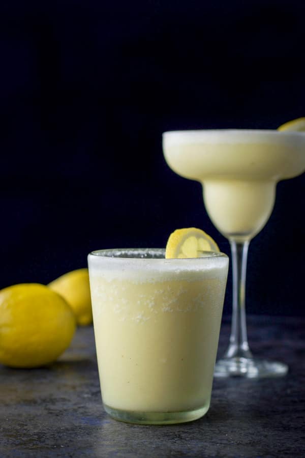 Vertical view of the lemon margarita in an old fashioned glass in front and in a classic margarita glass in back, next to two lemons