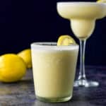 Double old fashioned glass filled with the margarita in front with the classic margarita glass in the background. There are lemons on the board